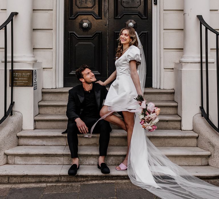 Bride in a short wedding dress with long wedding veil standing next to her husband dressed all in black sat on some steps in Mayfair 