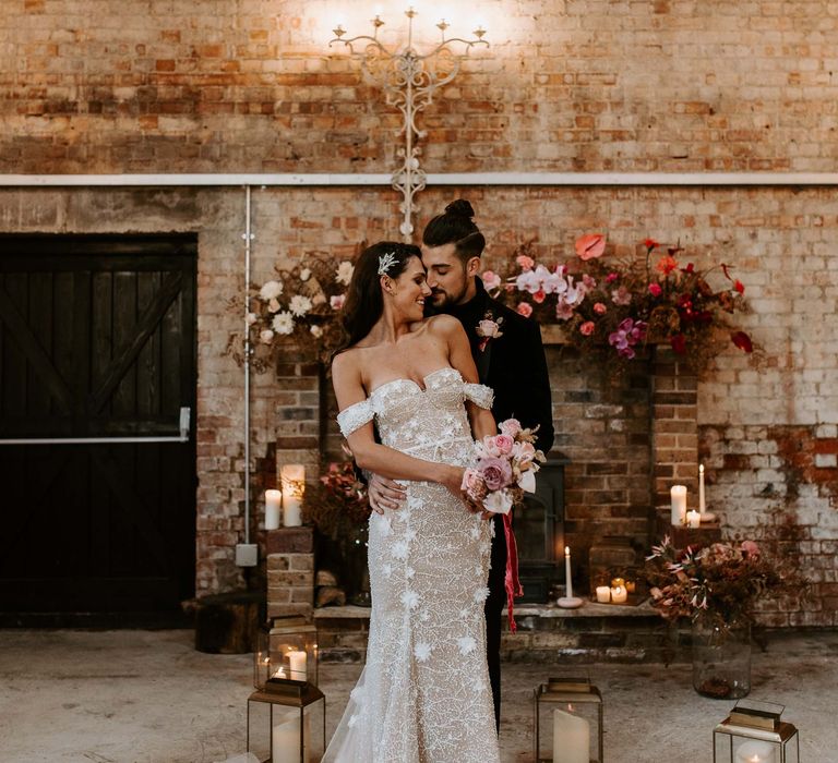 Bride in an off the shoulder wedding dress surrounded by black lanterns holding a small lilac and pink bouquet 