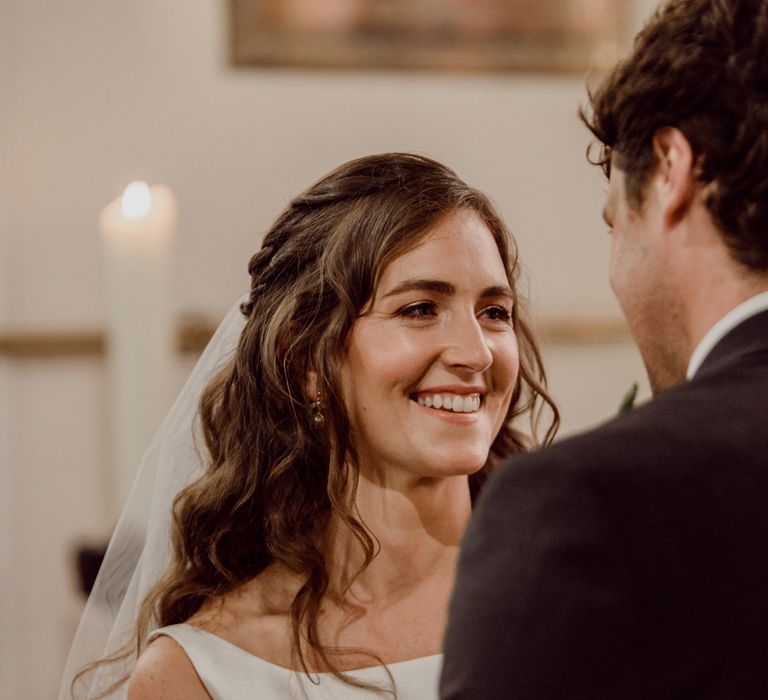 Smiling bride with brown curled hair and veil looks at groom in dark brown suit during church wedding ceremony