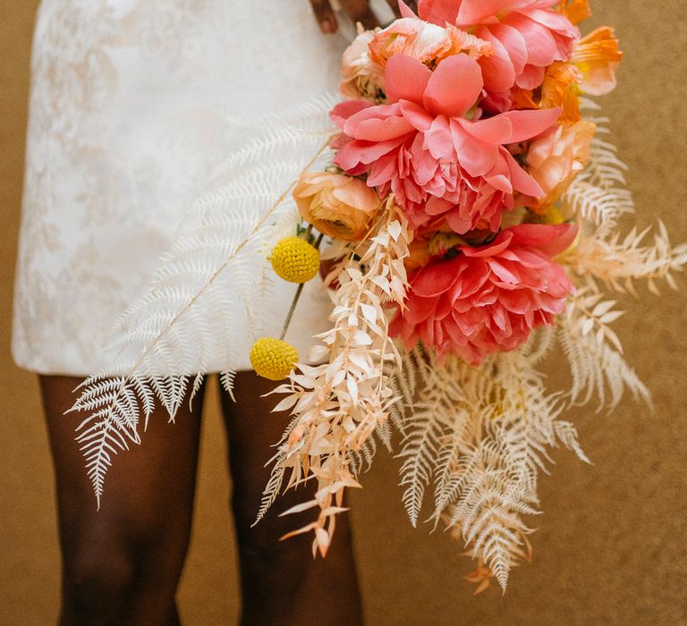 Bride wearing an embroidered mini skirt holding a bouquet of large pink dahlias, dried ferns and yellow craspedias