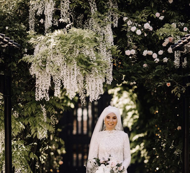 Bride stands under hanging wisteria in high neck and long sleeve 3D lace wedding dress with white orchid and rose wedding bouquet