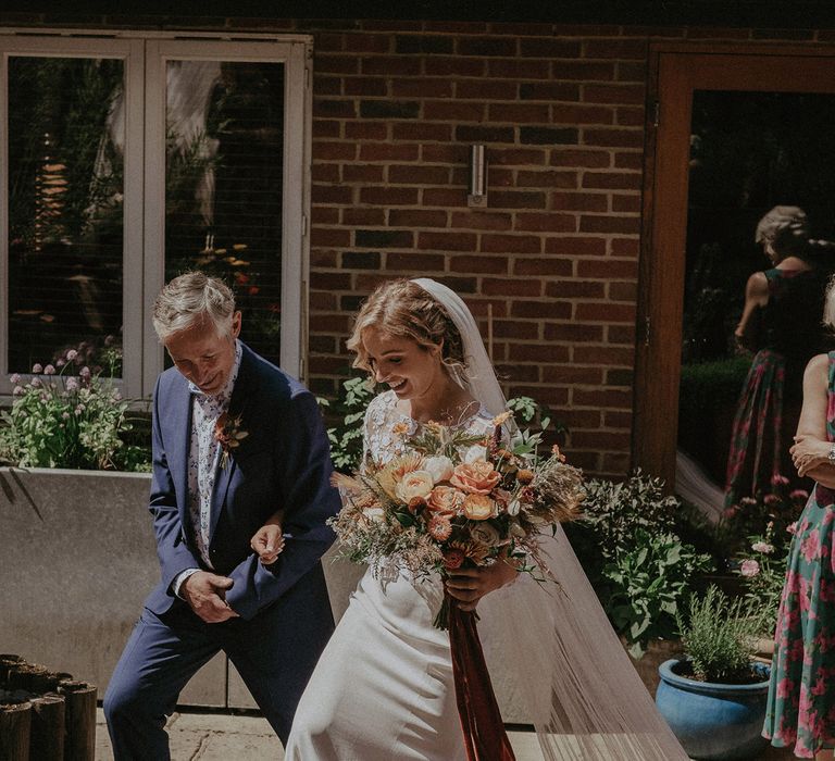 Bride in white Rime Arodaky wedding dress with mesh floral embroidered top and veil carries burnt orange bridal bouquet whilst walking arm-in-arm with father in blue suit up steps at garden wedding with burnt orange wedding theme