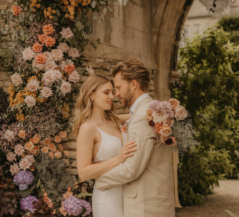 Bride with long wavy hair wearing a sparkly wedding dress embracing her groom in a beige suit with her pastel wedding bouquet over his shoulder 