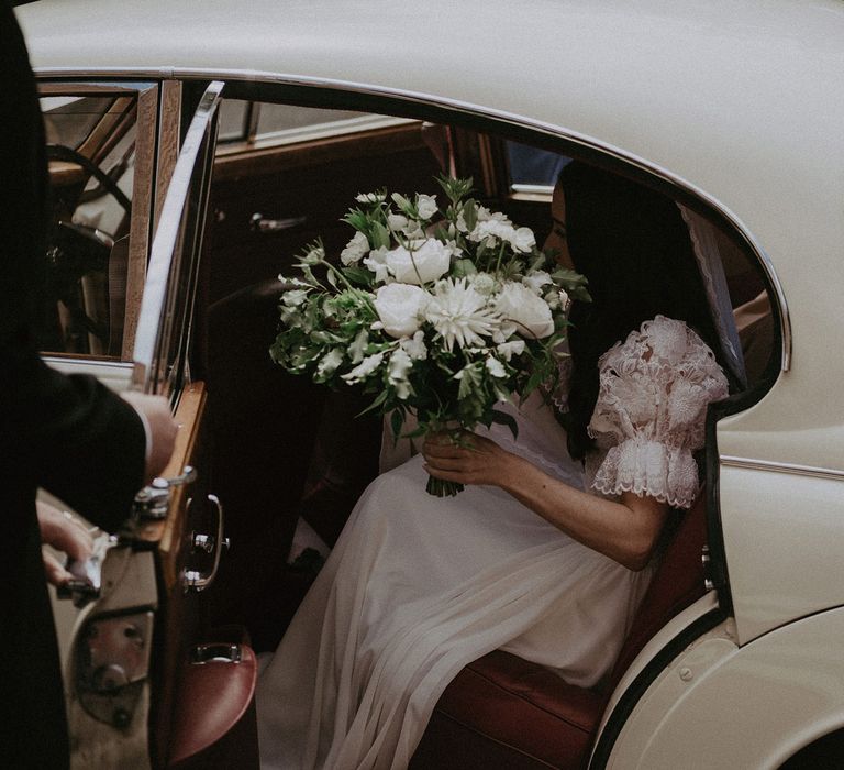 Bride in lace puffed sleeve Daalarna wedding dress holding white and green bridal bouquet gets out go white classic car before church wedding ceremony in Surrey