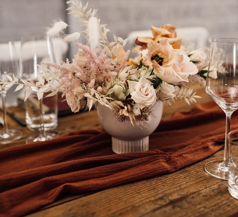 Small bowl of mixed fresh and dried florals in pink bowl on burgundy table runner for boho wedding breakfast with long wooden tables at summer wedding in Dorset