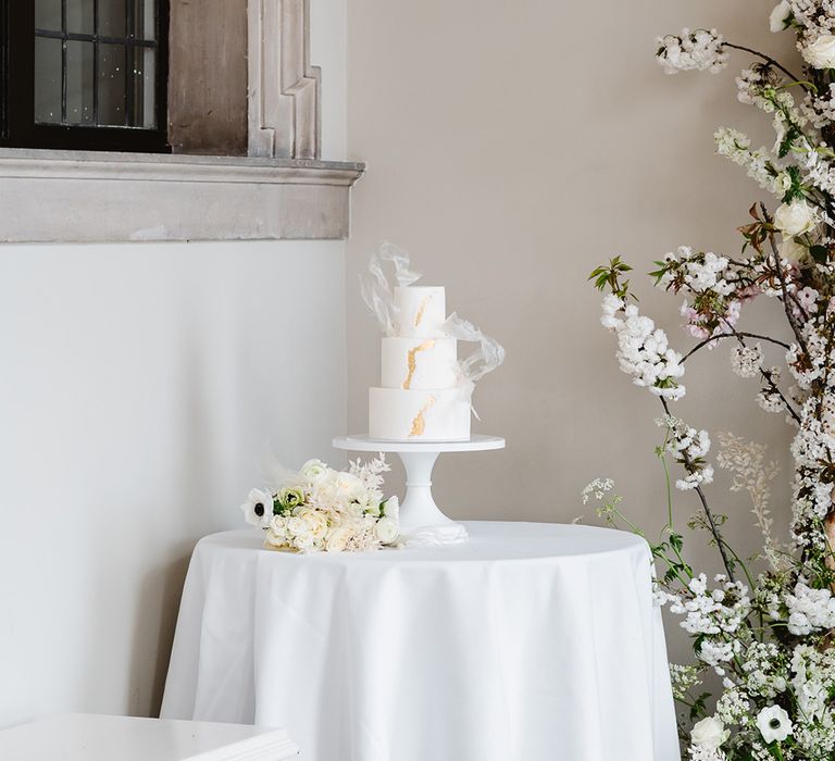 A white wedding cake at a black and white wedding. The cake has three tiers and gold leaf detailing. 