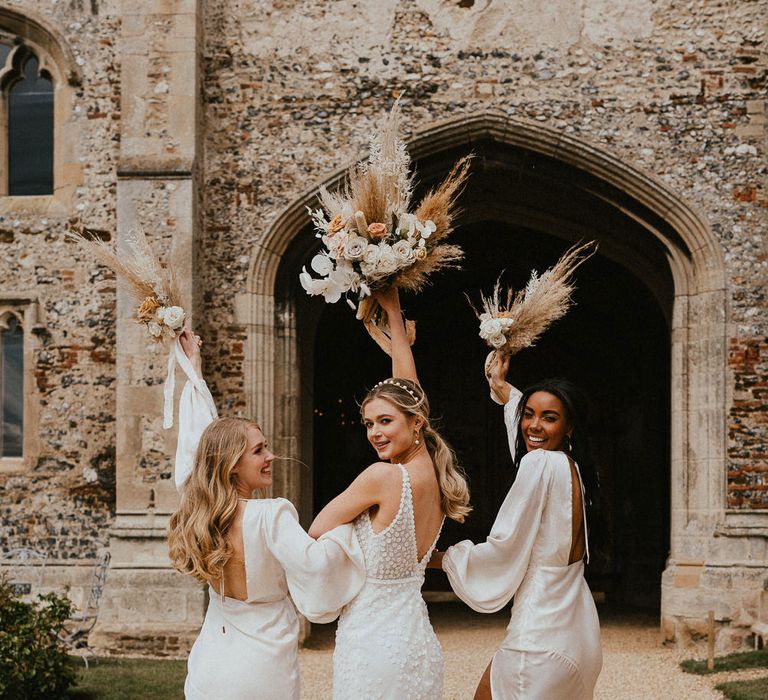 Bride in an appliqué wedding dress and bridesmaids in neutral satin wrap dresses holding their bouquets in the air 