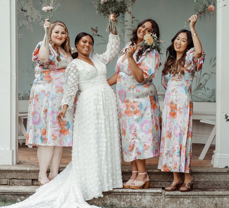 Bride in white long mesh sleeve spotted wedding dress stands with bridesmaids in multicoloured floral dresses all holding mixed bouquets in the air outside Victorian summerhouse at Wasing Park wedding