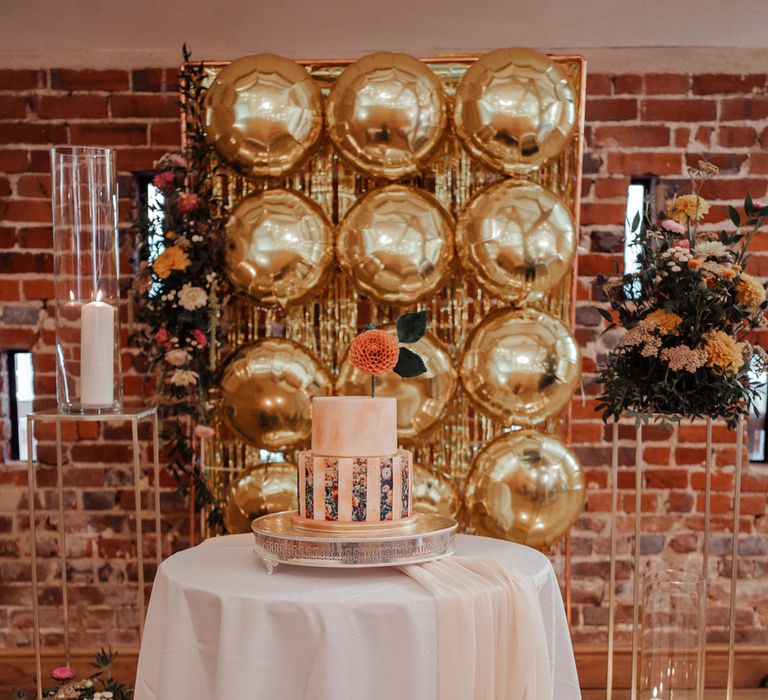  Blush pink marble effect two tier wedding cake with floral photo detailing and orange flower on top on round table with white table cloth in front of gold balloon wall surrounded by florals and candles