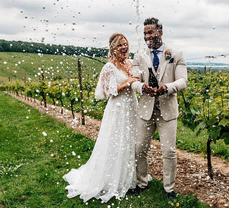 Bride in a lace dress and groom in beige suit popping champagne at Little Wold Vineyard 