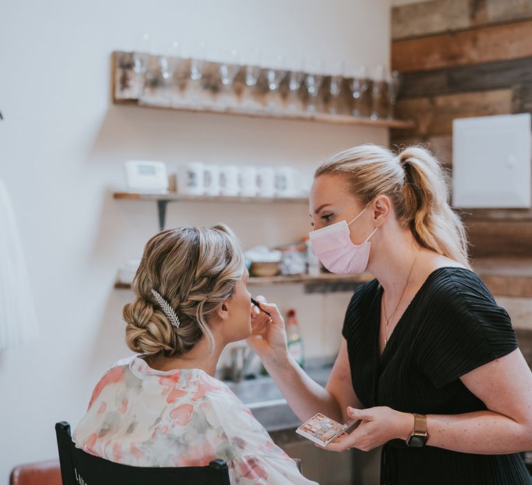 Bride with plaited bun and leaf hair pin sits having her make up done by woman in black jumpsuit and pink face mask before Tythe Barn wedding with barn wedding flowers