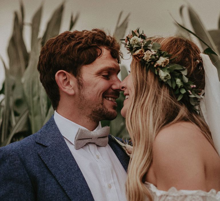 Bride in Grace Loves Lace wedding dress, veil and flower crown smiles with groom in blue suit jacket and sage green bow tie as they stand in corn field at Isle of Wight wedding with macrame wedding decor