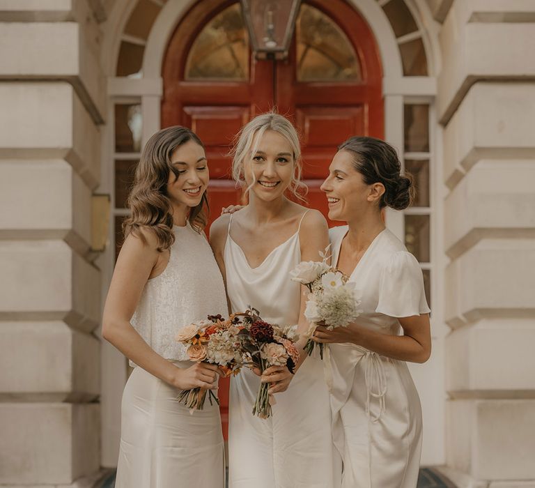 Bridesmaids in Constellation Are white satin dresses and jumpsuits standing on the steps at Clerkenwell House holding orange flower bouquets 