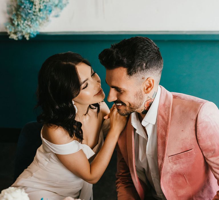 Groom in a gold shoulder wedding dress leaning in to kiss her groom in a pink velvet dinner jacket 