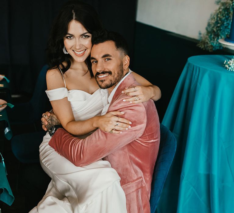 Bride in a cold shoulder wedding dress sitting on her grooms lap in a pink velvet jacket 