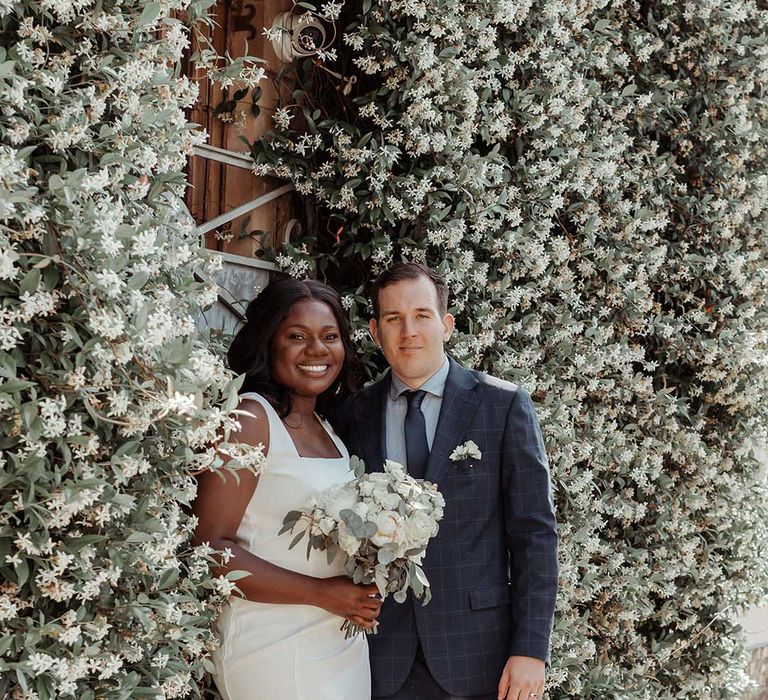 Bride & groom stand beside white floral wall on their wedding day in Chelsea