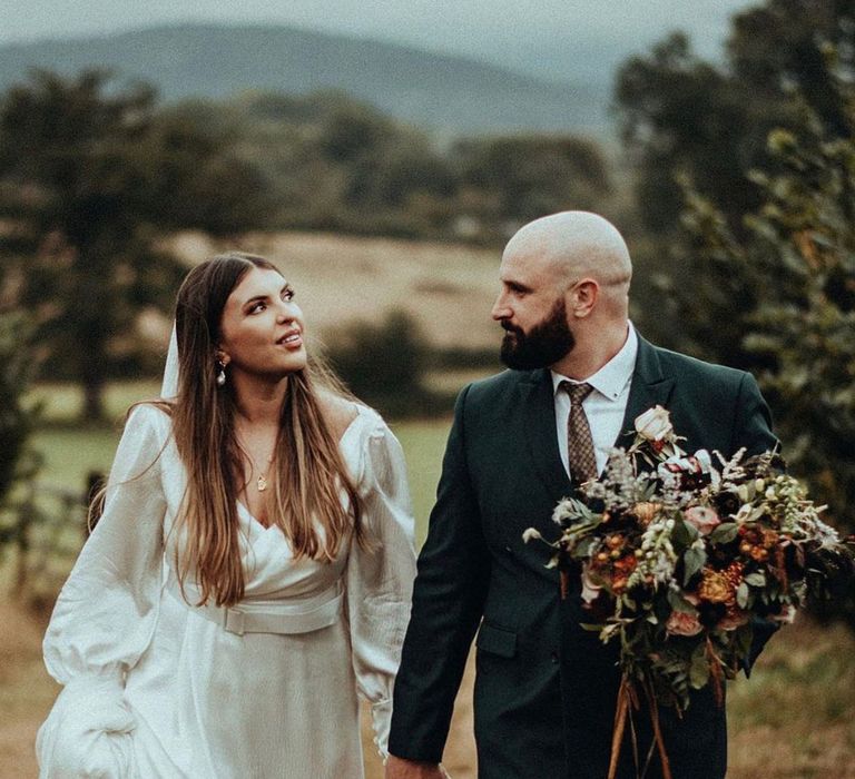 Bride & groom walk together across fields as the groom holds floral bouquet