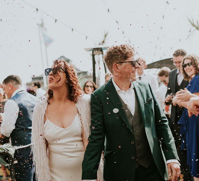 Bride & groom walk hand in hand through confetti as bride playfully sticks her tongue out to wedding guest