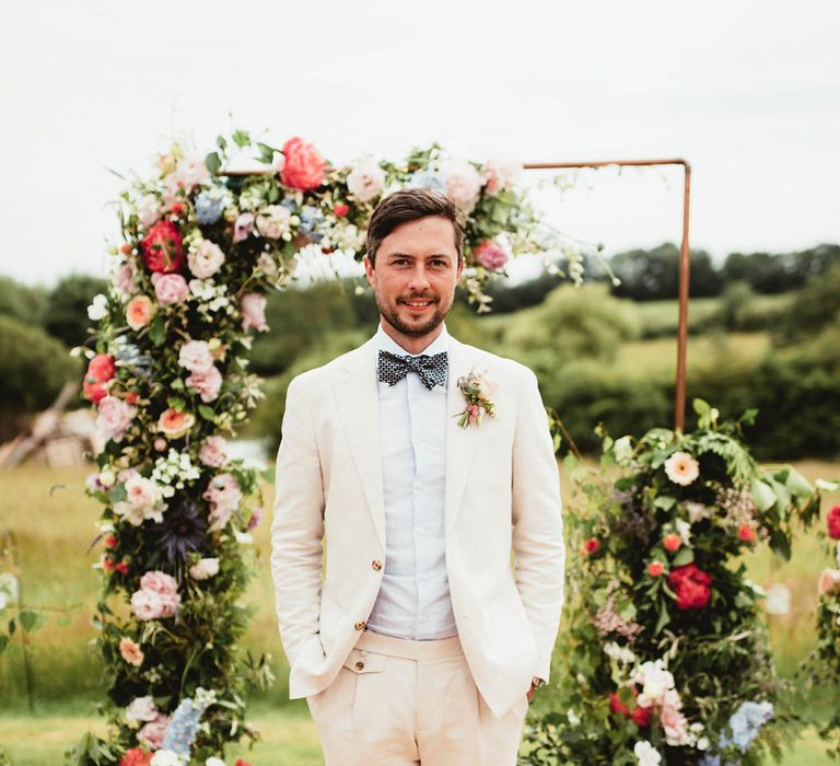 Groom stands in front of floral archway on his wedding day