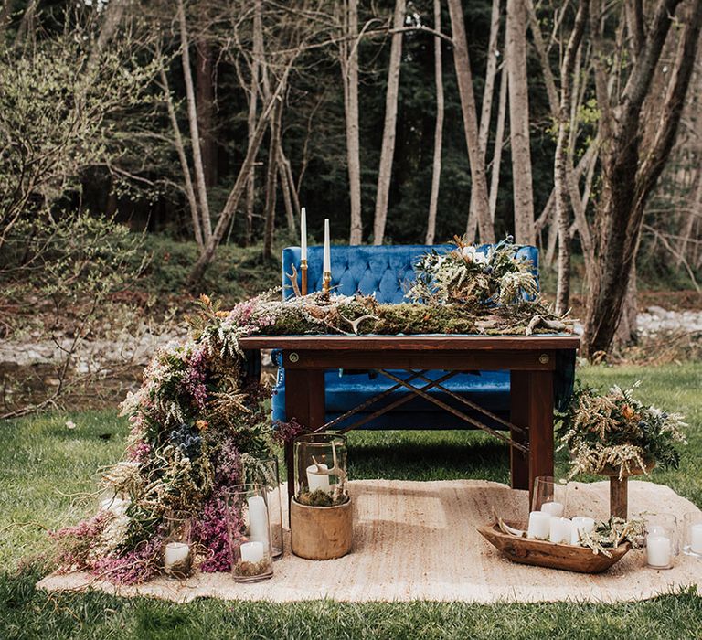 Outdoor sweetheart table with plush blue velvet love seat and trailing wild flower arrangement 