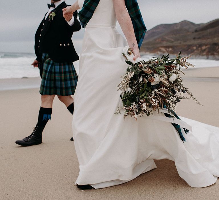 Bride holding her boho bouquet with anemones, wildflowers and foliage by her side as she walks along the beach 