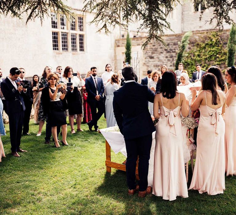 Bridesmaid in blue suit and bridesmaids in baby pink satin bridesmaid dresses with bow back take part in sugar ceremony outside at Elmore Court