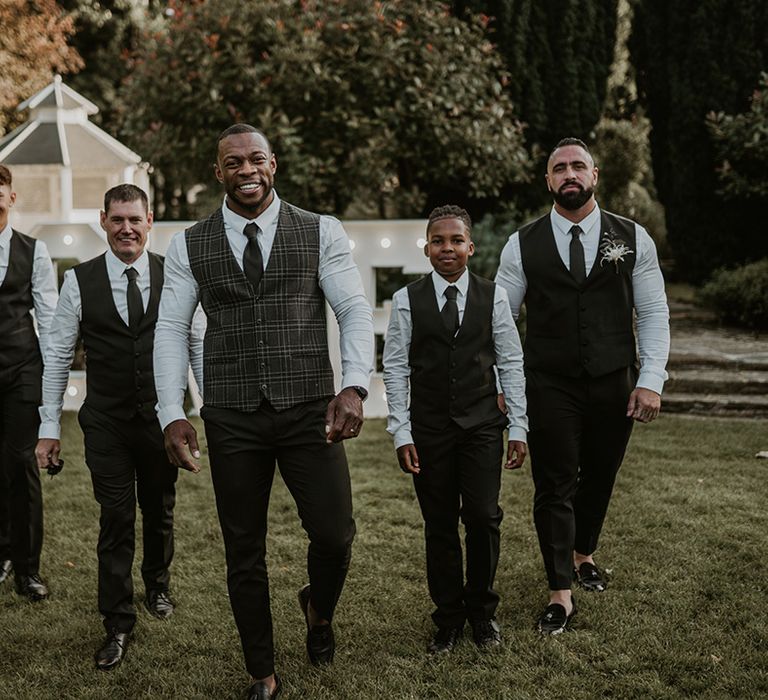 Groom smiles as he wears tartan waistcoat complete with green trousers and walks with his groomsmen 