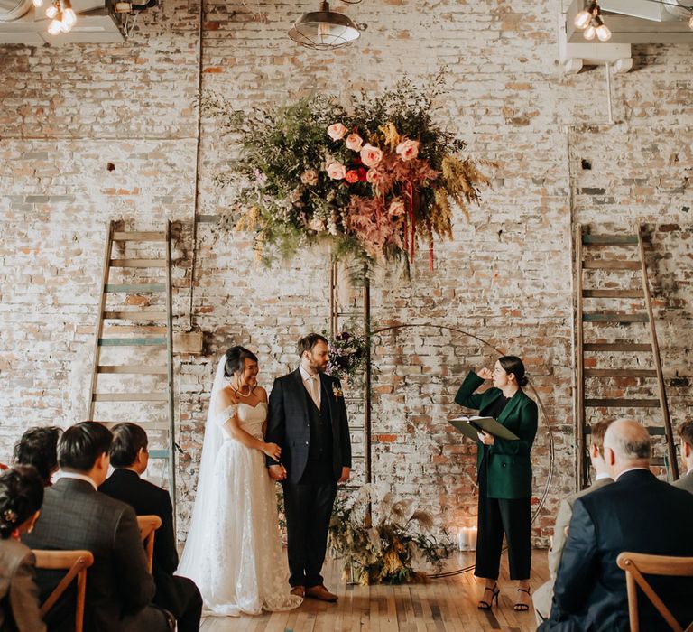 Bride in lace wedding dress and groom in blue check suit stand under mixed pink, yellow and green flower cloud with foliage and florals during wedding ceremony at Scale in Liverpool