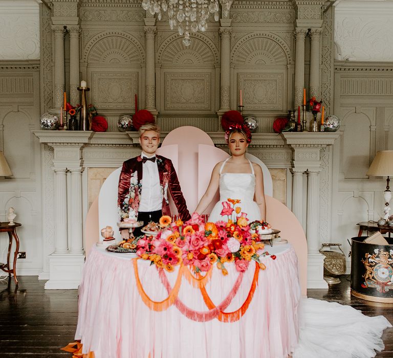 Two brides in a Burgundy velvet blazer and tulle skirt wedding dress holding hands in front of a  pink geometric backdrop, red, pink and orange flower arrangement and food by Baba Ganoush 