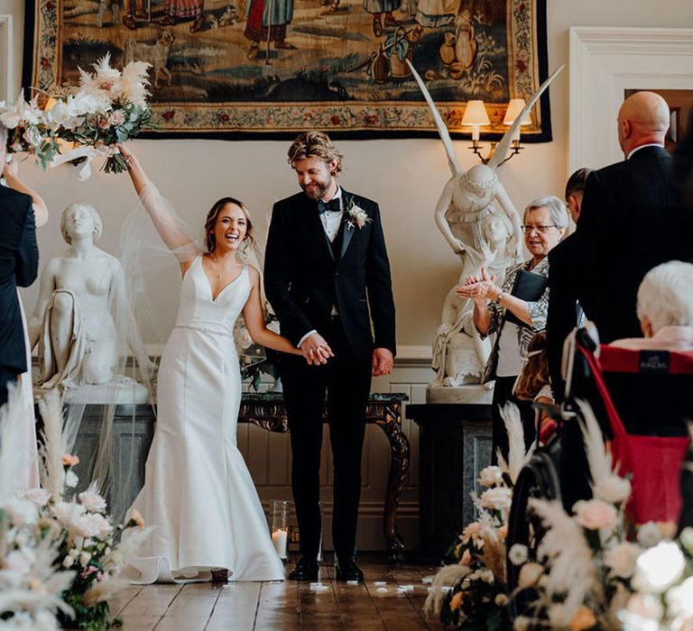 Bride in fishtail wedding dress and neutral bouquet of flowers and groom in three-piece black tuxedo