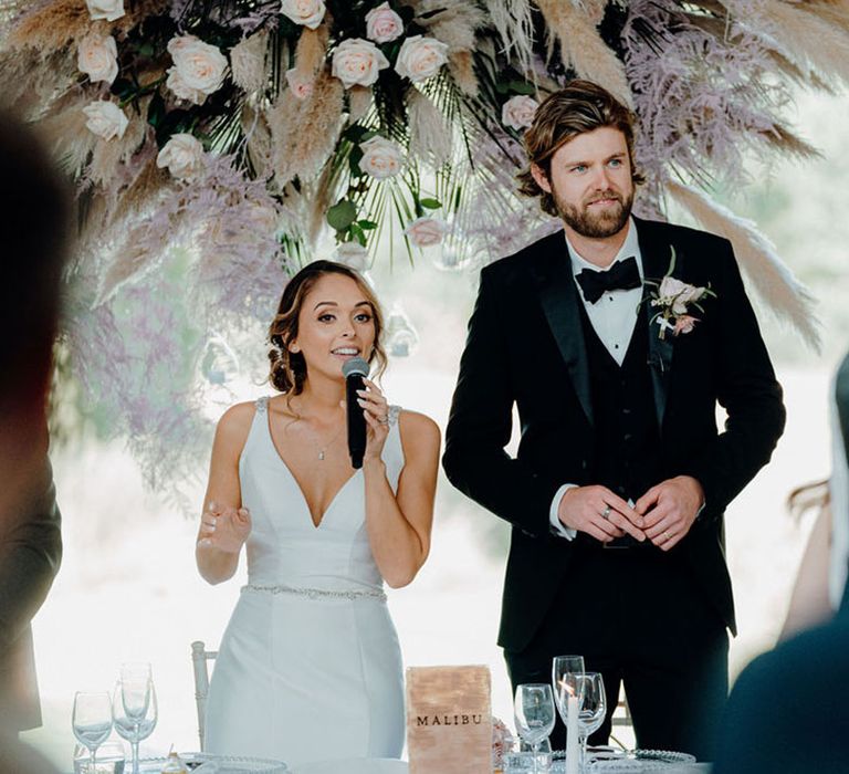 Bride in a fitted wedding dress giving her speech under a flower installation at Elmore Court 