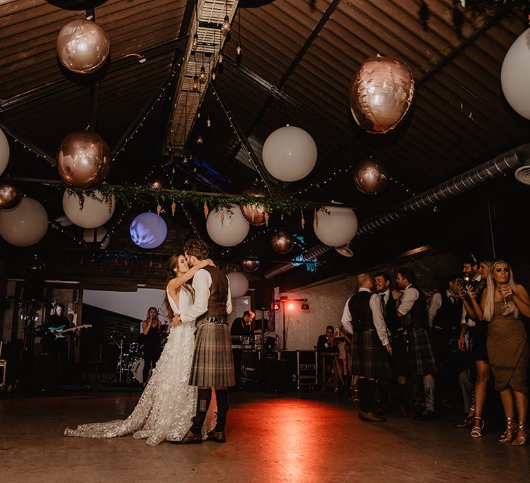 Bride and groom dance under large white and rose gold balloon decoration with groomsmen in kilts in the background