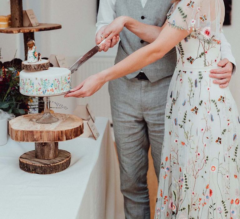 Bride in embroidered floral wedding dress with groom in grey waistcoat cut their white flowery wedding cake on tree trunk stand