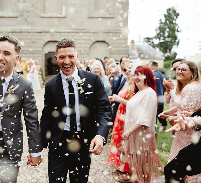 Smiling and happy grooms walk through confetti after their wedding ceremony in black and grey suits and dried flower buttonholes