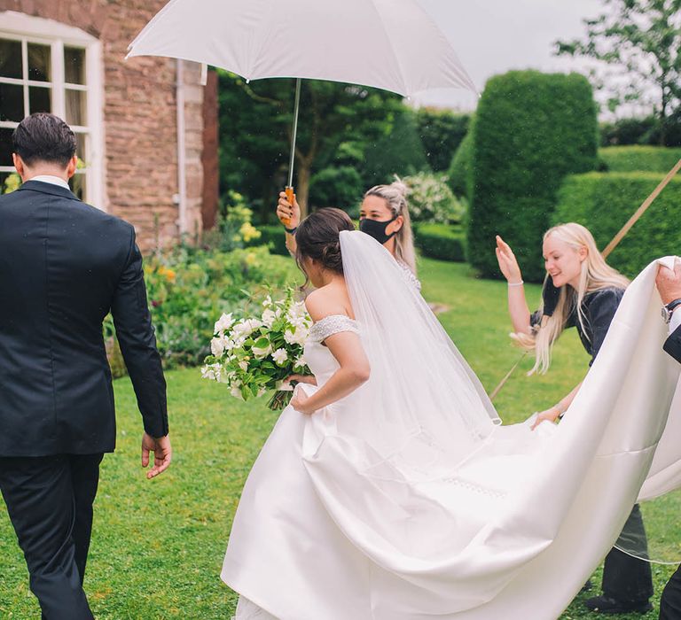 Bride and groom walk to venue under an umbrella with bride holding white flower and greenery wedding bouquet