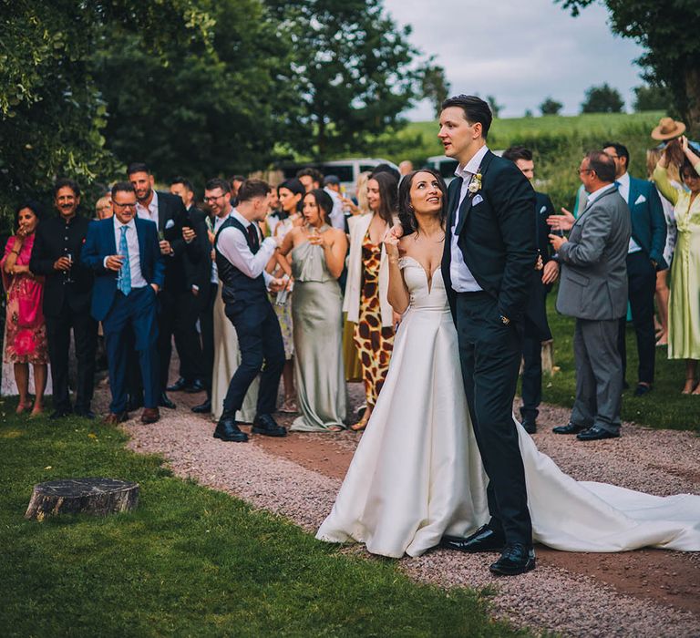 Groom in black tie has his arm around bride in sparkly off the shoulder wedding dress with guests socialising 