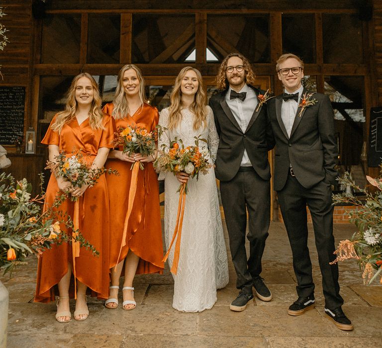 Bride in lace wedding dress with bridesmaids in burnt orange satin wrap dresses and groom and groomsmen in matching black tie