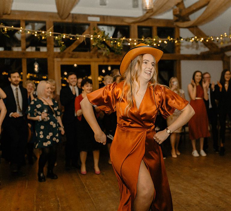 Bridesmaid in Rewritten Bridesmaids burnt orange satin wrap dress with matching orange cowboy hat for line dance entertainment 