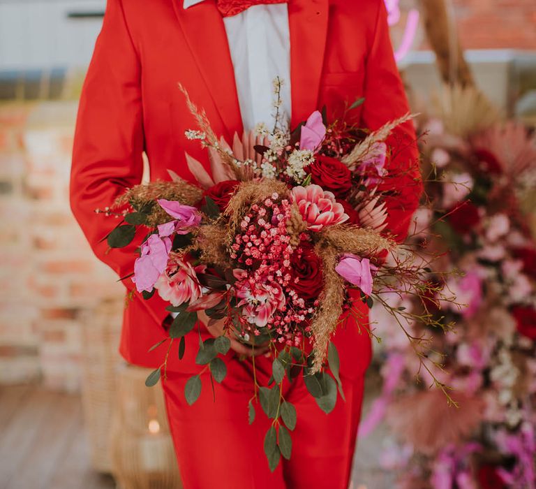 Groom in a red suit and bow tie holding a pink and red wedding bouquet at rustic barn wedding 