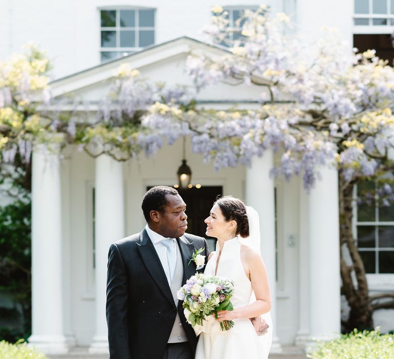 Groom in black morning suit jacket with bride in Jesus Peiro wedding dress outside Pembroke Lodge venue with purple wisteria 