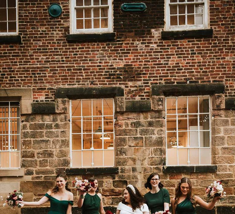 Bridesmaids in different styles of forest green dresses with bride in short wedding dress at industrial wedding venue 