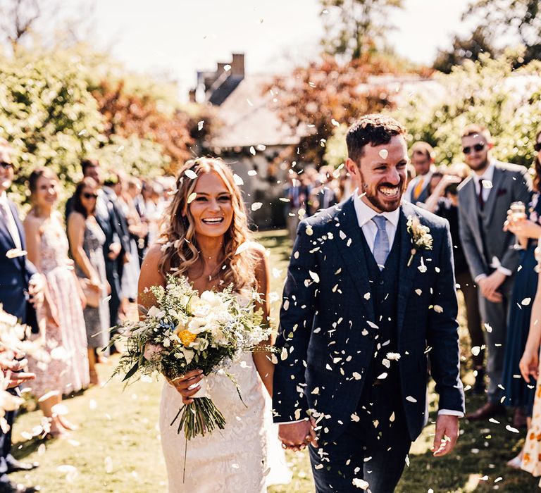 Bride carrying yellow bouquet holding hands with groom in blue suit have white confetti exit from ceremony 