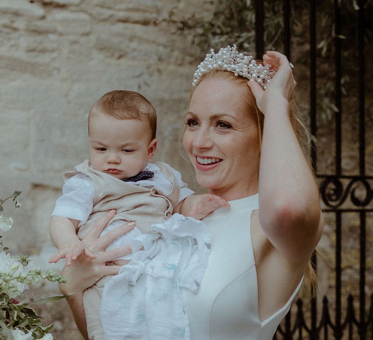 Bride adjusts her pearl headpiece as she holds a baby in her arms wearing beige trousers and waistcoat and a cute bow tie 