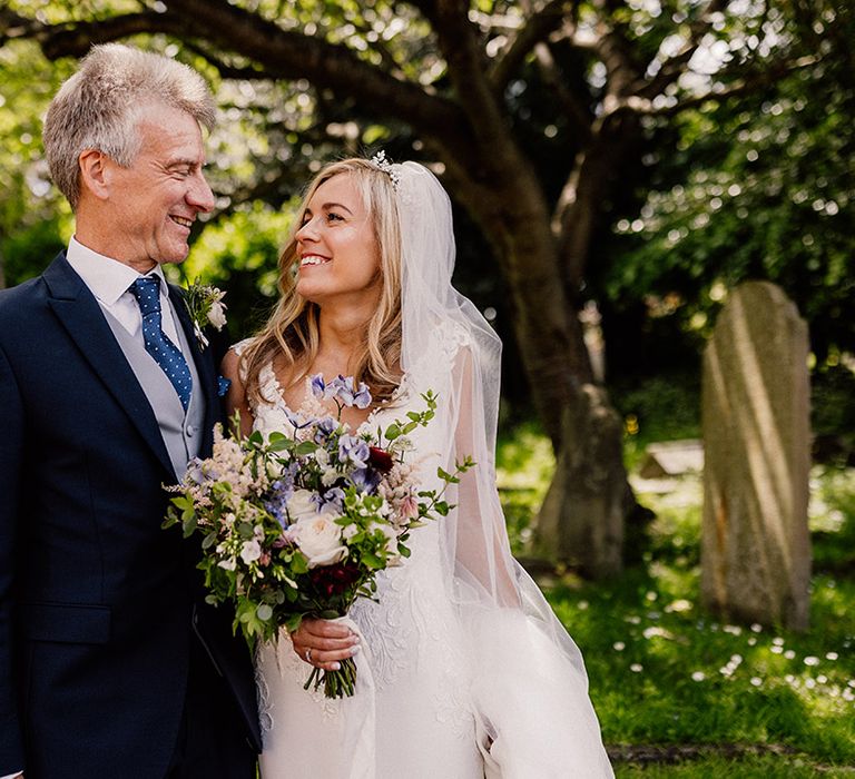 Bride in lace wedding dress with spring bouquet smiles at her father who wears a black suit with polka dot blue tie and handkerchief 