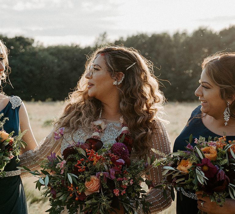 Bride holds colourful floral bouquet and looks toward her bridesmaids who wear navy blue bridesmaids gowns with embellishment to shoulder