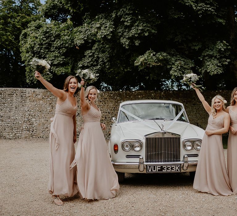 Bridesmaids in champagne pink dresses raise their matching white rose bouquets in the air as they stand next to the white wedding car 