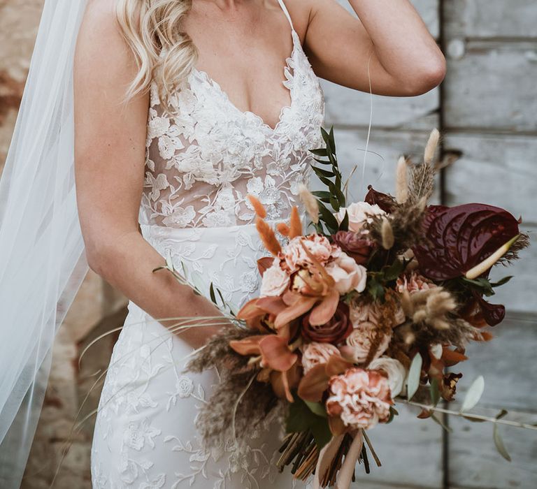 Bride wears romantic lace Emmy Mae Bridal wedding dress with thin straps and holding colourful floral bouquet 