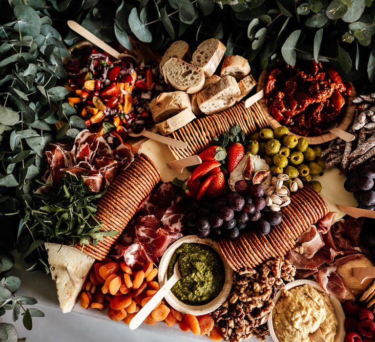 Colourful Spread grazing board complete with biscuits, hummus and pesto dips, tomatoes, olives, grapes, meats and manchego cheese  