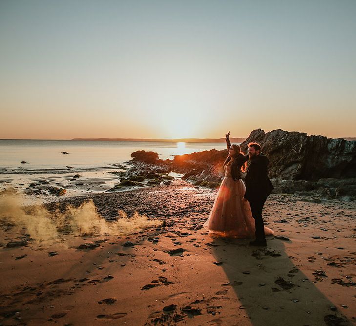 Bride & groom walk along beach during golden hour surrounded by colourful smoke bombs 