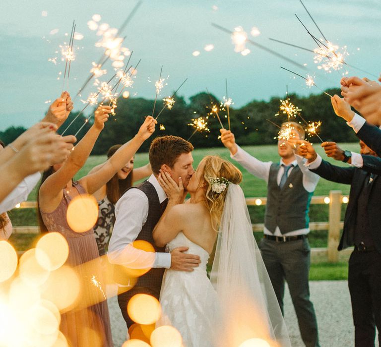 Bride in a strapless wedding dress kisses the groom as their guests circle around them waving sparklers 